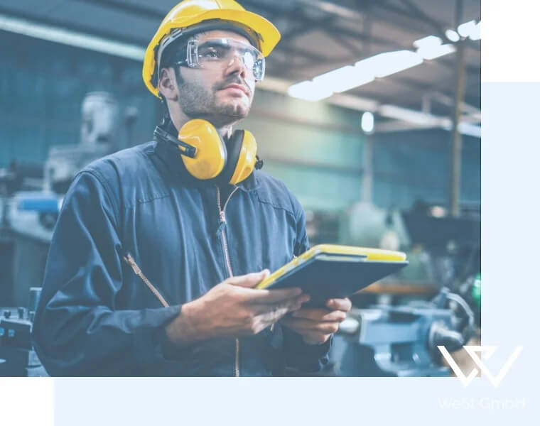 Mann in Arbeitskleidung mit Helm, Hörschutz und Schutzbrille trägt ein Tablet in der Hand.